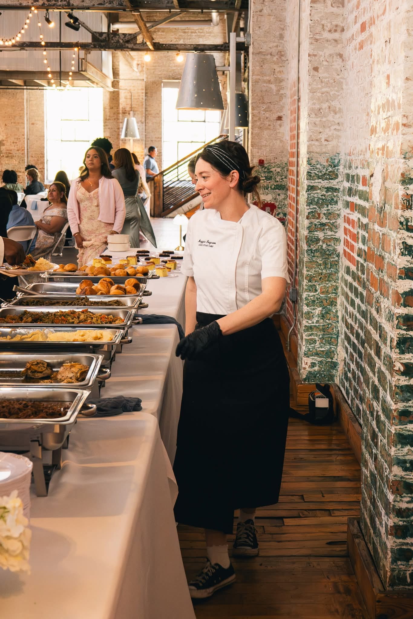 Chef Maggie Ingram beside a full catering buffet in a brick warehouse venue with string lights