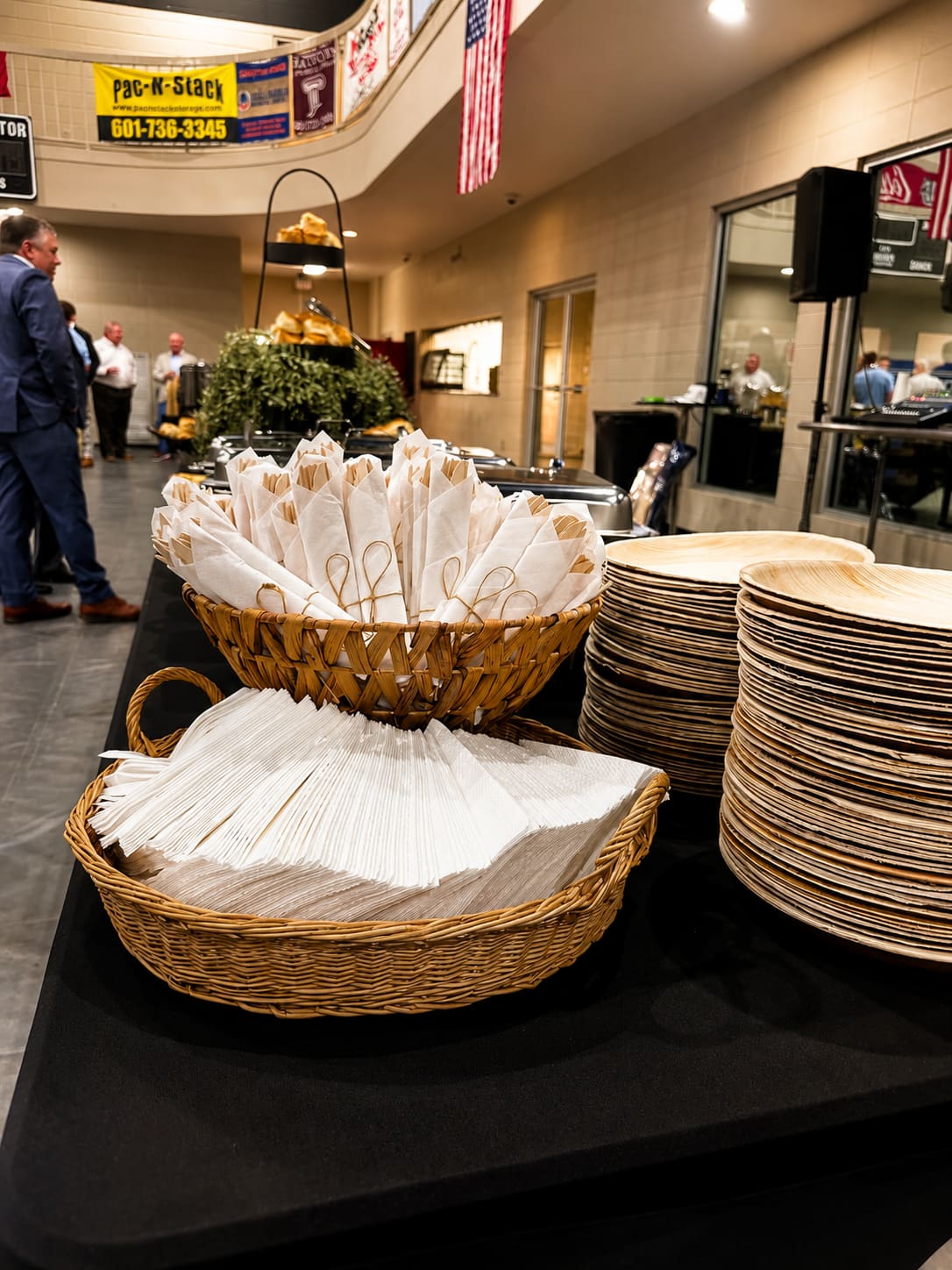Rolled napkins in woven baskets and stacked palm-leaf plates set out for a catered event